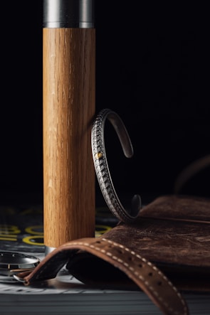 A wooden cylindrical object with a metallic top stands upright. A detailed silver bracelet with a textured design hangs on the wood. In the foreground, a brown leather strap with stitching is partially visible, alongside what appears to be the edge of a watch case. A leather-bound book or notebook is positioned in the background. The setting is dimly lit, creating a rich contrast among the textures and materials.