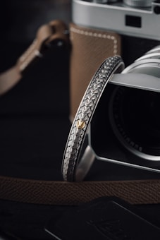 A close-up of a camera with a brown leather strap and a detailed, intricately engraved metallic ring attached. The camera features a black lens and silver body, highlighting precision craftsmanship. The focus is on the textured design and the gold accent on the ring, with a soft and dark background enhancing the product's elegance.