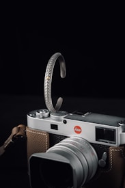 A vintage style camera with a silver body and brown leather grip features prominently. Draped over the top of the camera is an intricately designed silver bracelet with detailed etchings and a small gold accent.