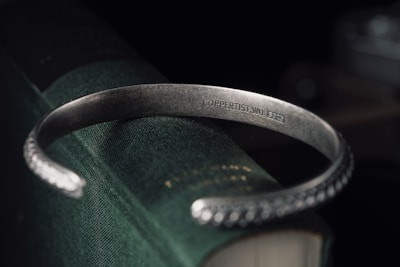 A metallic bracelet engraved with 'COPPERTIST.WU 5925' is draped over the corner of a closed, textured dark green book. The bracelet has a simple, polished design with intricate patterns on its ends.