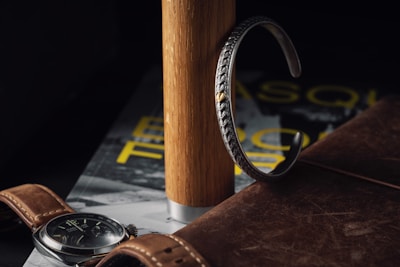A leather watch with a brown strap and a dark face is placed on top of a magazine. Beside it is a silver bracelet with intricate designs, leaning against a wooden cylinder. The atmosphere is cozy and sophisticated with a dark, moody background.