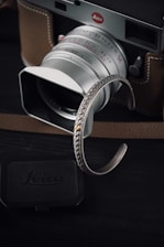 A silver camera with a brown leather case is prominently featured. In the foreground, a detailed silver bracelet with intricate patterns adds a touch of elegance. The camera is classic with visible brand insignia, and a lens cap is placed nearby. The overall setting is dark, highlighting the metallic and leather surfaces.