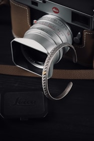 A silver camera with a brown leather case is prominently featured. In the foreground, a detailed silver bracelet with intricate patterns adds a touch of elegance. The camera is classic with visible brand insignia, and a lens cap is placed nearby. The overall setting is dark, highlighting the metallic and leather surfaces.