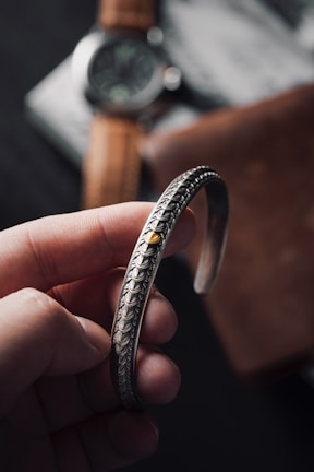 A hand holding a detailed, intricately designed metallic bracelet with a central gold accent. In the background, a blurred watch with a leather strap and part of a wooden item are visible.