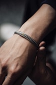 A close-up of a person's wrist wearing an intricately designed metallic bracelet with a small gold accent. The skin is illuminated by soft lighting, highlighting the texture of both the bracelet and the skin.