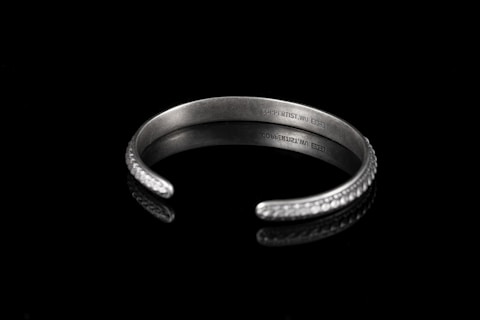 A silver bracelet with embossed patterns resting on a glossy black surface. The inside of the bracelet is engraved with the text 'COPPERTIST.WU' and a series of numbers. The bracelet has an open-ended design and the surface reflects light, enhancing its lustrous appearance.