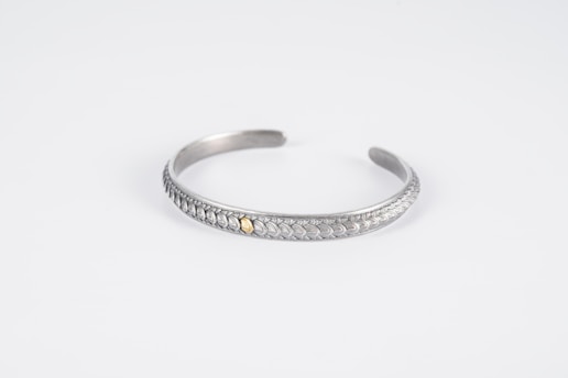 A silver bracelet with intricate leaf-like patterns and a small gold accent in the center is displayed on a plain white background. The bracelet is curved into an open, circular shape with a smooth, polished finish.