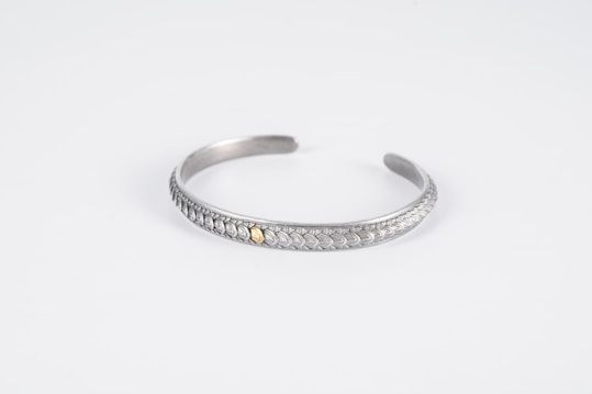 A silver bracelet with intricate leaf-like patterns and a small gold accent in the center is displayed on a plain white background. The bracelet is curved into an open, circular shape with a smooth, polished finish.