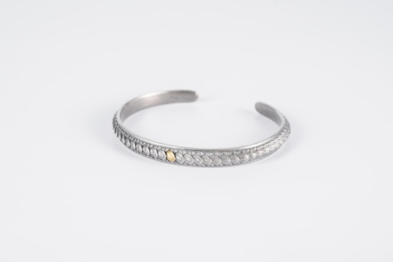 A silver bracelet with intricate leaf-like patterns and a small gold accent in the center is displayed on a plain white background. The bracelet is curved into an open, circular shape with a smooth, polished finish.