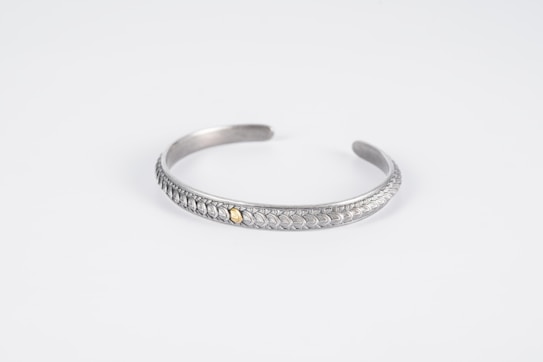 A silver bracelet with intricate leaf-like patterns and a small gold accent in the center is displayed on a plain white background. The bracelet is curved into an open, circular shape with a smooth, polished finish.