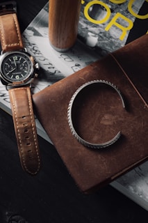 A stylish wristwatch with a brown leather strap rests on a stack of books. Beside it, a silver bracelet lies on a brown leather-bound notebook. The scene conveys a sense of sophistication and classic style, with a background featuring a book with yellow text and a wooden cylindrical object.