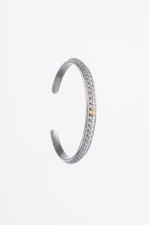 A silver bracelet with intricate decorative patterns along its exterior. The bracelet has an open-ended design and a small bronze or golden accent near the opening.