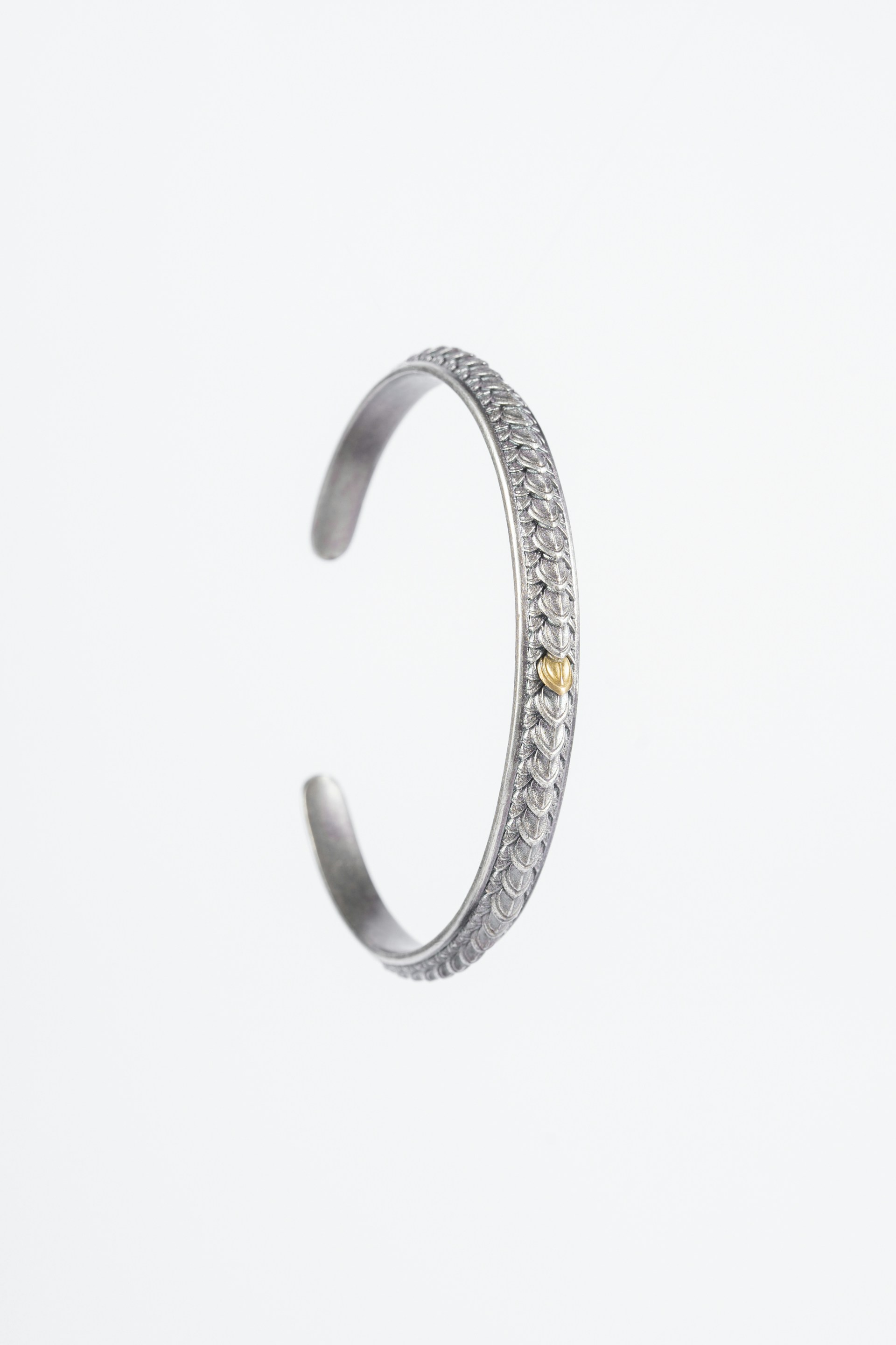 A silver bracelet with intricate decorative patterns along its exterior. The bracelet has an open-ended design and a small bronze or golden accent near the opening.