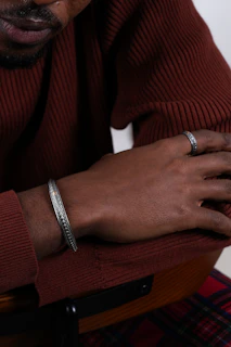 A close-up of layered accessories on a woman’s wrist and hand, featuring earth-toned bracelets and rings against a #a68e7d sweater