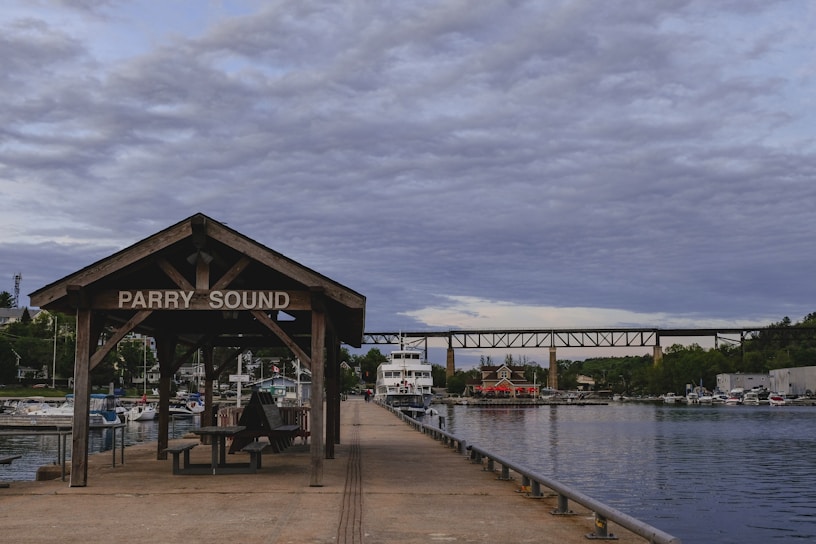 A wooden shelter with a sign reading 'Parry Sound' stands on a pier extending into calm water. Various boats are docked along the pier, and a large white boat is visible further down. In the background, there is a bridge spanning across the water and several buildings with greenery around them. The sky is filled with dense, layered clouds.