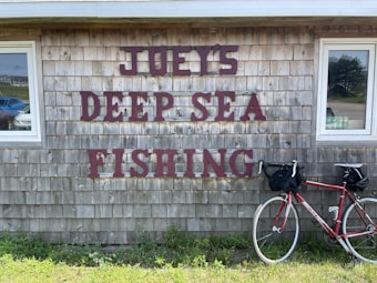 A rustic shingle-clad building with large, bold letters reading 'JOEY'S DEEP SEA FISHING' mounted on the wall. A red bicycle is parked against the building, positioned beneath the sign. There are two windows on either side of the text, reflecting the outdoor surroundings, including some parked vehicles and greenery.