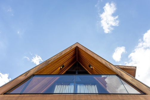 A modern A-frame wooden cabin is viewed from below against a backdrop of a clear blue sky scattered with a few white clouds. The structure features large glass windows reflecting the sky, and the wooden panels are warmly lit by outdoor lights.