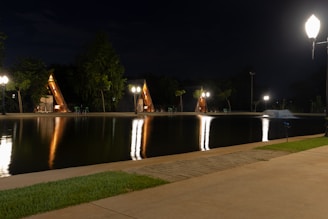 Small A-frame cabins are illuminated at night with street lamps reflecting on a serene pond. The scene is surrounded by trees, and the ground is paved with both cement and grass.