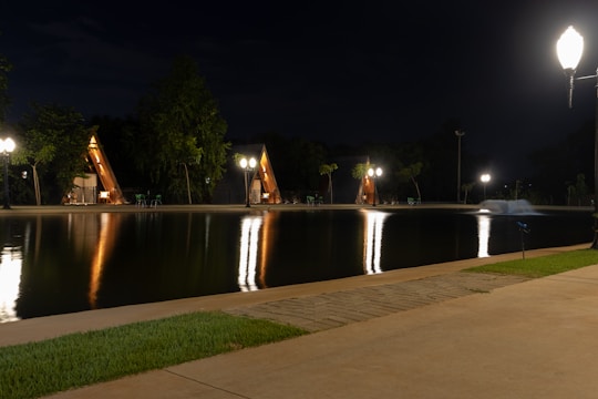 Small A-frame cabins are illuminated at night with street lamps reflecting on a serene pond. The scene is surrounded by trees, and the ground is paved with both cement and grass.