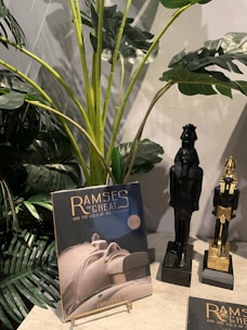 A book titled 'Ramses the Great and the Gold of the Pharaohs' is displayed on a stand next to two black and gold Egyptian statues. In the background, there are large green tropical plants with broad leaves.