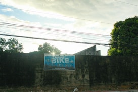 A large, weathered wall with a blue and white sign advertising bicycles for sale is prominently displayed in an outdoor setting. Various power lines run horizontally across the upper part of the scene, and lush green trees and foliage appear in the background. The sky is partly cloudy with sunlight peeking through.