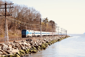 A passenger train travels along a track situated near a large body of water, bordered by rocky terrain. Dense, leafless trees line the opposite side of the track, with a series of utility poles running parallel to the rails.