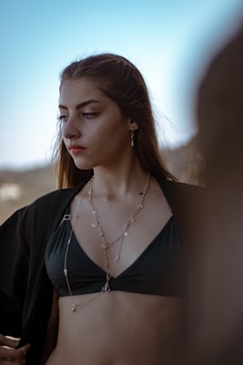 A young woman with long hair looks to the side, wearing a black bikini top and a delicate necklace with multiple charms. She is outside, with a blurred natural background that suggests an outdoor setting. The ambient light highlights her facial features and the texture of her clothing.