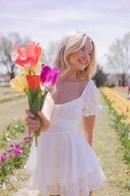A person with long blonde hair is smiling while holding a colorful bouquet of tulips. The scene is set outdoors in a vibrant flower field with rows of blooming tulips under a clear sky.
