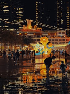 A vibrant, illuminated urban scene featuring a festive, nighttime gathering. Colorful structures and light displays create a lively atmosphere. People are walking and interacting, with reflections visible on the wet ground, adding to the dynamic environment.