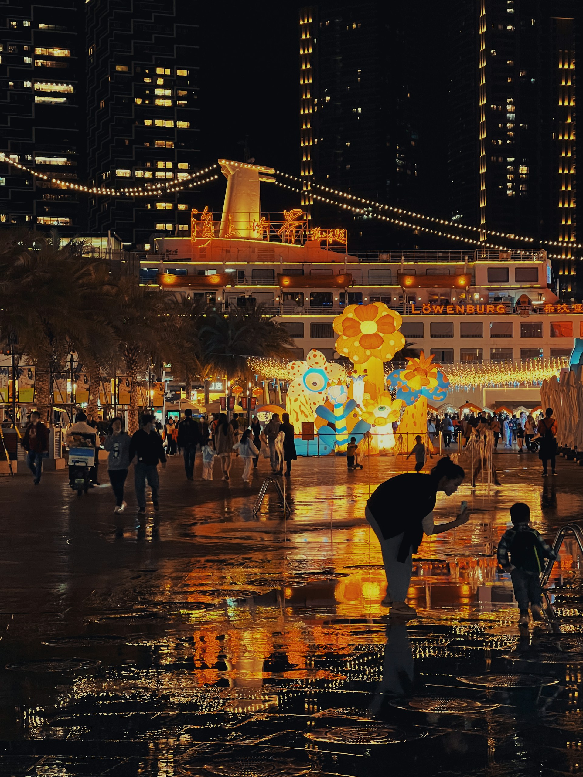 A vibrant, illuminated urban scene featuring a festive, nighttime gathering. Colorful structures and light displays create a lively atmosphere. People are walking and interacting, with reflections visible on the wet ground, adding to the dynamic environment.
