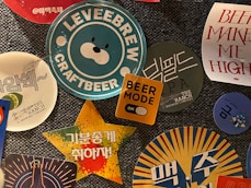 A collection of colorful beer-themed stickers and coasters, featuring various logos, text, and graphics. The central image displays a blue and white sticker with a bear face and the text 'LEVEEBREW CRAFTBEER'. Surrounding it are other designs including a yellow square with 'BEER MODE' and an orange toggle, a star-shaped sticker with Korean text on a vibrant background, and additional circular coasters with brewery logos and phrases.