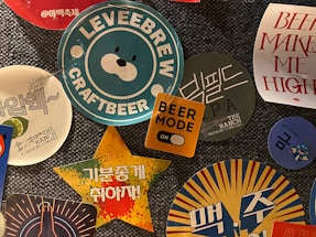A collection of colorful beer-themed stickers and coasters, featuring various logos, text, and graphics. The central image displays a blue and white sticker with a bear face and the text 'LEVEEBREW CRAFTBEER'. Surrounding it are other designs including a yellow square with 'BEER MODE' and an orange toggle, a star-shaped sticker with Korean text on a vibrant background, and additional circular coasters with brewery logos and phrases.