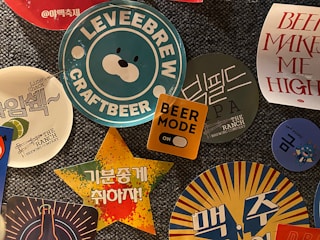 A collection of colorful beer-themed stickers and coasters, featuring various logos, text, and graphics. The central image displays a blue and white sticker with a bear face and the text 'LEVEEBREW CRAFTBEER'. Surrounding it are other designs including a yellow square with 'BEER MODE' and an orange toggle, a star-shaped sticker with Korean text on a vibrant background, and additional circular coasters with brewery logos and phrases.