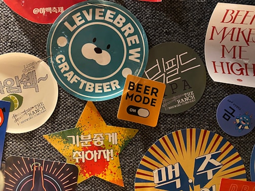 A collection of colorful beer-themed stickers and coasters, featuring various logos, text, and graphics. The central image displays a blue and white sticker with a bear face and the text 'LEVEEBREW CRAFTBEER'. Surrounding it are other designs including a yellow square with 'BEER MODE' and an orange toggle, a star-shaped sticker with Korean text on a vibrant background, and additional circular coasters with brewery logos and phrases.