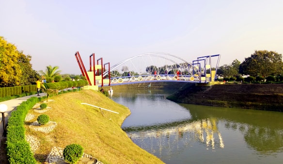 A landscaped park with a futuristic, colorful bridge spanning over a calm river. The bridge features geometric shapes and vibrant colors, such as blue and red. On the left, trimmed hedges and a walkway can be seen, with a person walking. The surrounding area includes a variety of trees and shrubs, contributing to the serene environment.