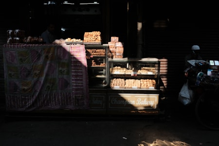 A street bakery stall displaying various baked goods, including bread and pastries, inside a glass case. The setup is partially covered by a colorful patterned cloth. Sunlight filters through, casting shadows and highlighting the baked items. A person is partially visible in the background.