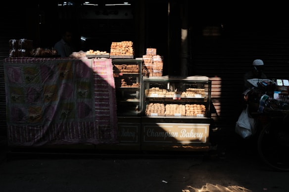 A street bakery stall displaying various baked goods, including bread and pastries, inside a glass case. The setup is partially covered by a colorful patterned cloth. Sunlight filters through, casting shadows and highlighting the baked items. A person is partially visible in the background.