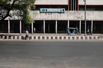 A city street scene with a man walking across the road, a parked blue auto-rickshaw nearby, and a building in the background with a sign reading 'Ravindra Tubes LTD'. The building appears old and has a series of columns supporting it. Another person is seen standing near the building, and a large tree provides shade to part of the scene.