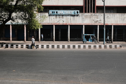 A city street scene with a man walking across the road, a parked blue auto-rickshaw nearby, and a building in the background with a sign reading 'Ravindra Tubes LTD'. The building appears old and has a series of columns supporting it. Another person is seen standing near the building, and a large tree provides shade to part of the scene.