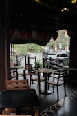 A cozy outdoor cafe area with wooden tables and chairs. Overhead, there are hanging lanterns, and the area is partially shaded. Potted plants and trees surround the space, creating a lush atmosphere. A black car is visible in the background, parked by the sidewalk with a view of an urban street lined with trees.