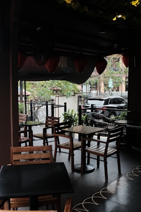 A cozy outdoor cafe area with wooden tables and chairs. Overhead, there are hanging lanterns, and the area is partially shaded. Potted plants and trees surround the space, creating a lush atmosphere. A black car is visible in the background, parked by the sidewalk with a view of an urban street lined with trees.