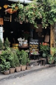 A quaint outdoor spot with lush greenery, displaying a variety of potted plants and flowers in baskets. A small, cozy storefront with Vietnamese signboards and hanging lanterns adds to the charm. There are stools and a table with utensils and a chalkboard promoting traditional herbal drinks. Ivy drapes from above, giving a calm, relaxed vibe.