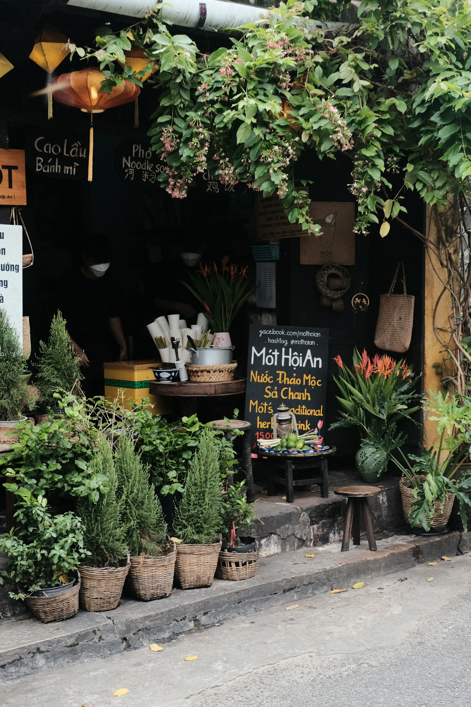 A quaint outdoor spot with lush greenery, displaying a variety of potted plants and flowers in baskets. A small, cozy storefront with Vietnamese signboards and hanging lanterns adds to the charm. There are stools and a table with utensils and a chalkboard promoting traditional herbal drinks. Ivy drapes from above, giving a calm, relaxed vibe.