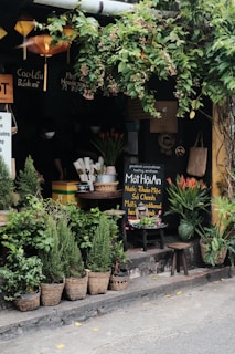 A quaint outdoor spot with lush greenery, displaying a variety of potted plants and flowers in baskets. A small, cozy storefront with Vietnamese signboards and hanging lanterns adds to the charm. There are stools and a table with utensils and a chalkboard promoting traditional herbal drinks. Ivy drapes from above, giving a calm, relaxed vibe.