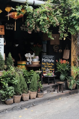 A quaint outdoor spot with lush greenery, displaying a variety of potted plants and flowers in baskets. A small, cozy storefront with Vietnamese signboards and hanging lanterns adds to the charm. There are stools and a table with utensils and a chalkboard promoting traditional herbal drinks. Ivy drapes from above, giving a calm, relaxed vibe.