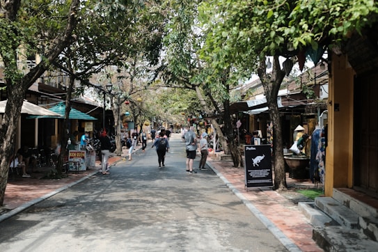 A lively street scene with people walking down a road lined with trees and shops. The street is shaded by tree branches and features small vendor stalls and outdoor seating areas. Colorful signs and decorations are visible, suggesting an atmosphere of a quaint market or shopping area.