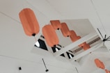 Several orange lanterns are hanging from the ceiling of a modern, white indoor space. The lanterns are irregularly spaced and aligned with the ceiling's architectural beams, with circular windows visible in the background.