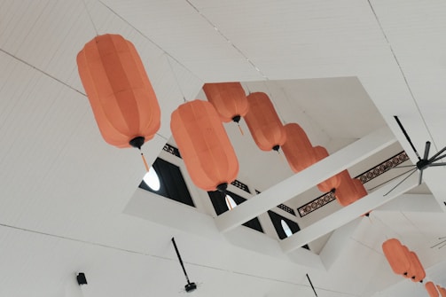 Several orange lanterns are hanging from the ceiling of a modern, white indoor space. The lanterns are irregularly spaced and aligned with the ceiling's architectural beams, with circular windows visible in the background.