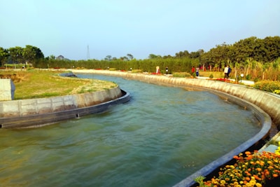 A curved canal flows through a landscaped area with lush greenery and trees in the background. Several people are walking along a path beside the canal, and vibrant flowers line the edge of the canal, adding color to the scene. The sky is clear with a soft blue hue, contributing to a serene atmosphere.