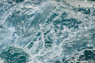 A close-up view of splashing water droplets, creating a dynamic and refreshing scene. The water appears in motion, with droplets suspended mid-air against a blurred aquatic background.